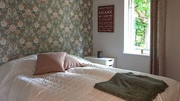 Un dormitorio con papel tapiz floral, una cama con cubrecama acolchado, una mesita de noche con lámpara y una ventana con cortinas.