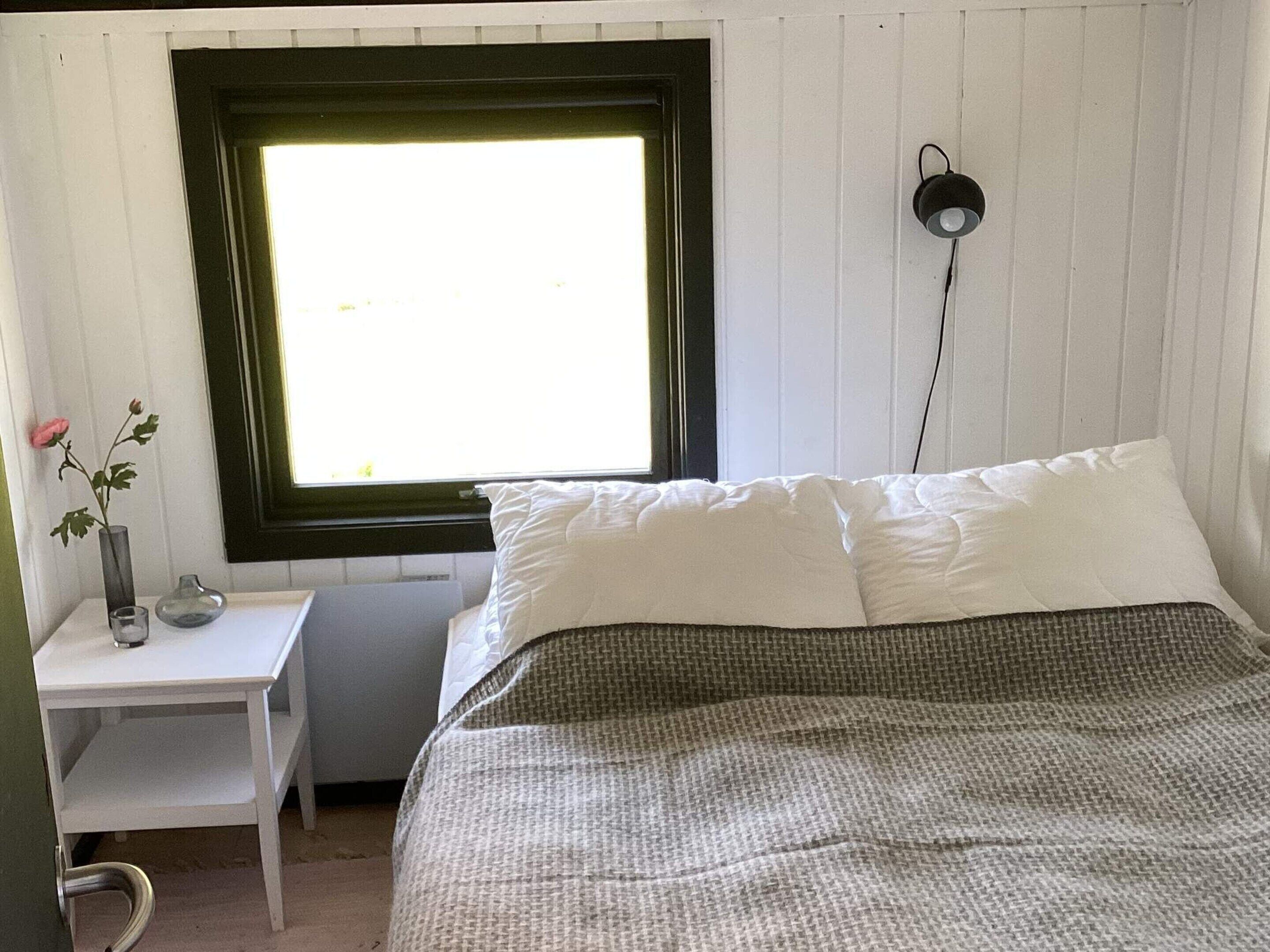 A bedroom with a bed, a bedside table with a vase and a glass, a window with a view, and a wall-mounted lamp.