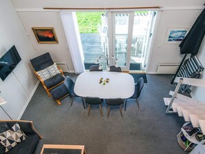 A room with a white table, four chairs, a TV, a sofa, and a window with a view of greenery.