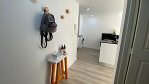 A hallway with a wooden floor, a white wall with a backpack and bottles on a small table, and a kitchen area in the background.