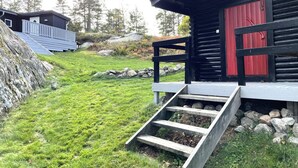 Une cabane avec un escalier en bois menant à une porte rouge, entourée d’une pelouse et d’arbres.