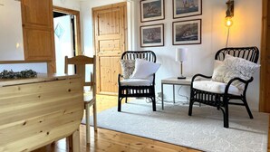 Une pièce chaleureuse avec un plancher en bois, une table à manger, deux chaises noires, une petite table blanche avec une lampe, et des tableaux marins encadrés accrochés au mur.