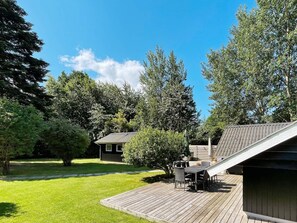 En træterrasse med et bord og stole, omgivet af en græsplæne og træer.