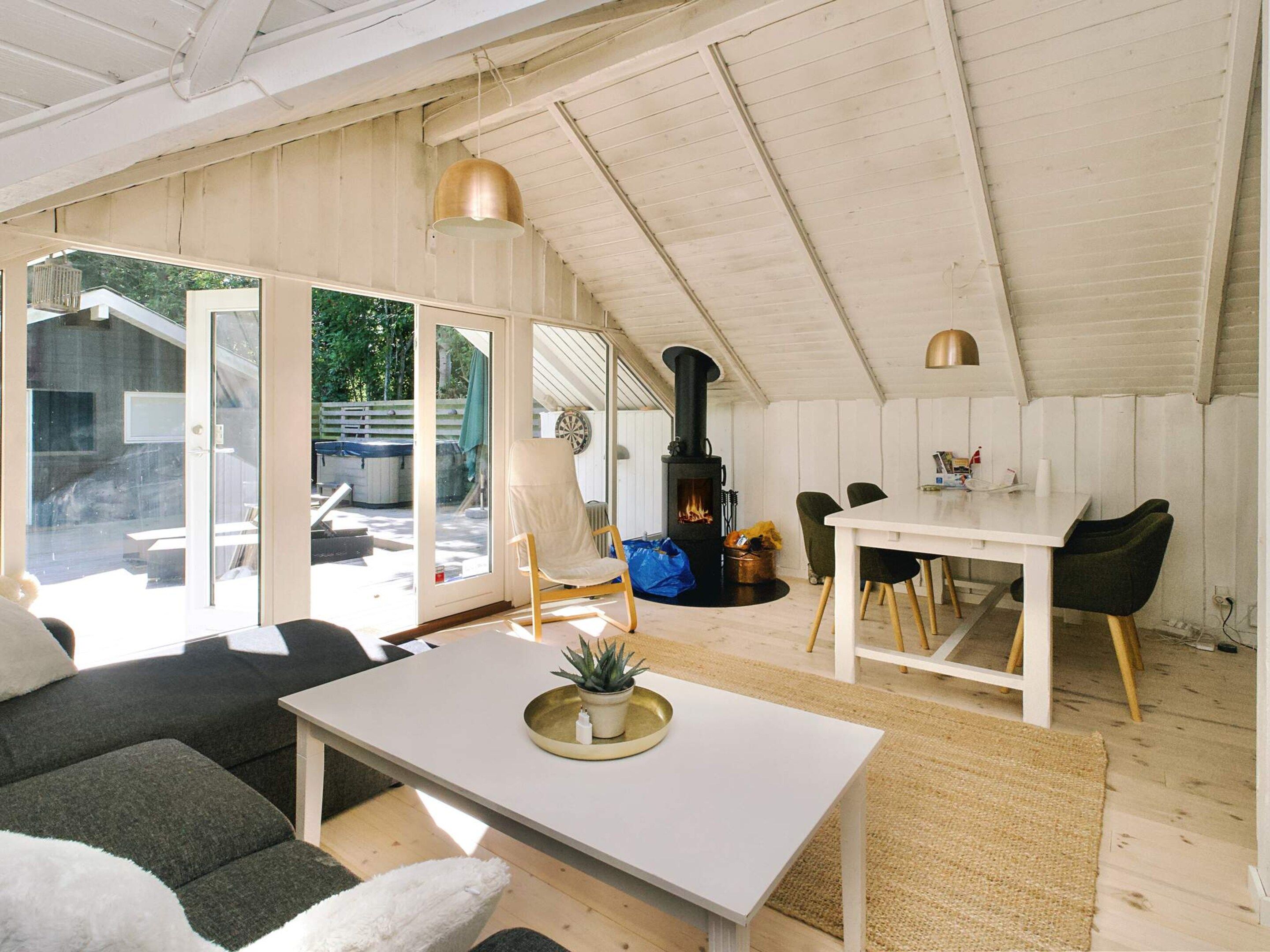 A cozy, well-lit room with a white ceiling, a black wood-burning stove, a dining table with chairs, and a sofa.