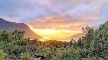 A mountainous landscape with a sunset, dense greenery, and a small building in the foreground.