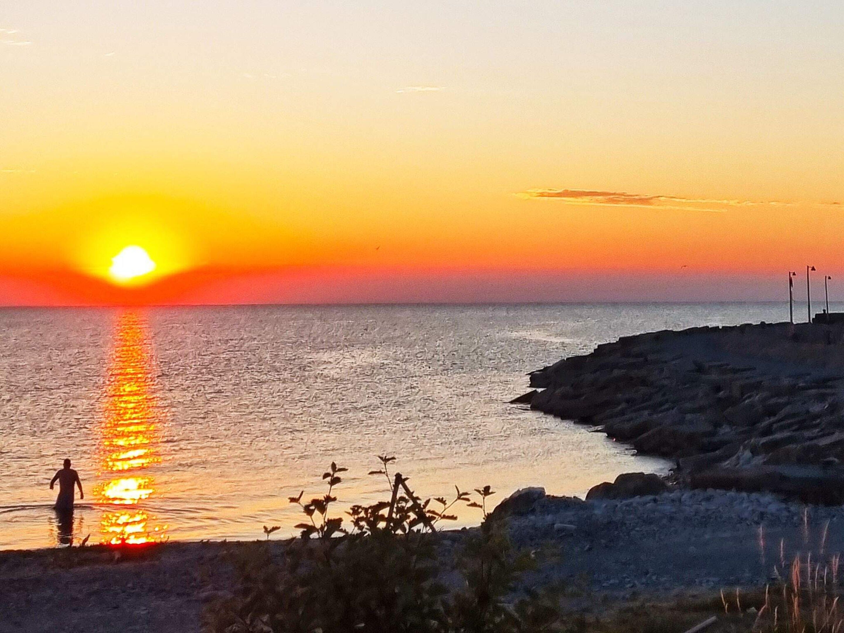 A sunset over a body of water with a rocky shoreline and a person standing in the water.