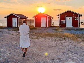 A person walking towards three wooden beach huts with doors, a clear sky, and the sun setting in the background.