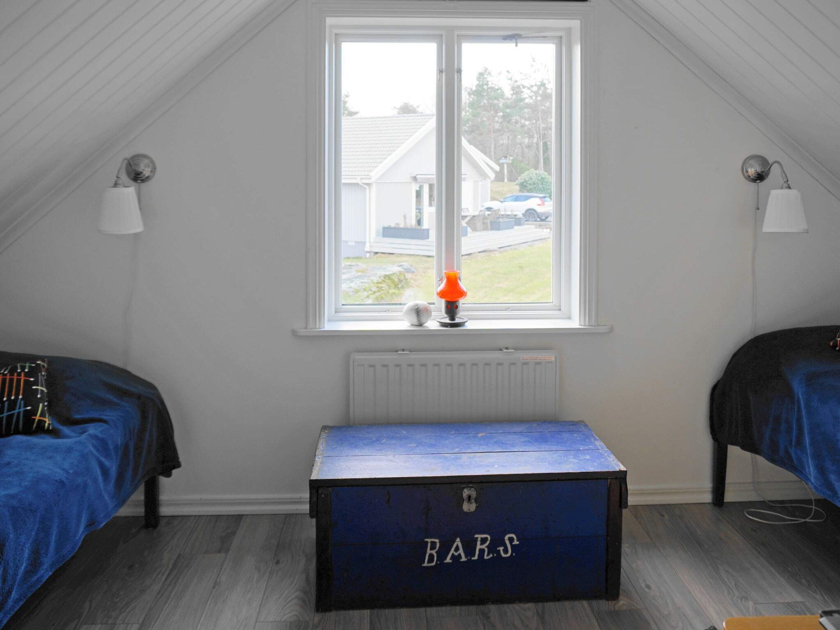 Une pièce avec un plafond en pente, une fenêtre donnant sur une maison, un radiateur et une commode bleue portant l’inscription « BARS ».