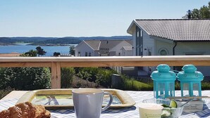 Une table dressée avec un croissant, une tasse, une assiette, un verre de lait et un pot de fromage frais, le tout placé sur un balcon donnant sur un lac et des maisons.