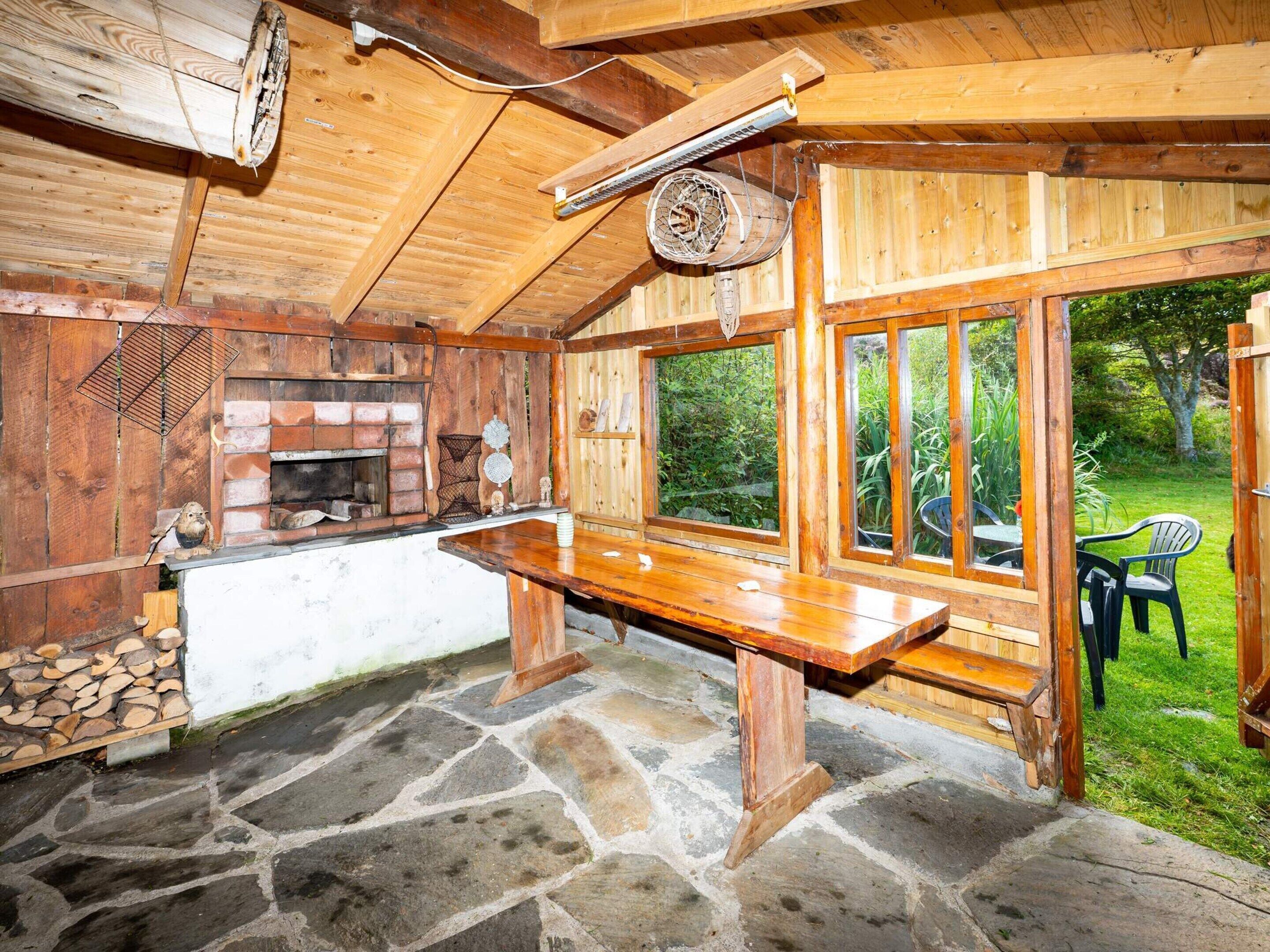 A rustic wooden cabin interior with a stone floor, a wooden bench, a brick fireplace, and large windows overlooking a garden.