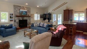 A spacious living room with a stone fireplace, a large blue sofa, a leather armchair, and a dining area with a high ceiling and natural light. - Torch Lake Cabin near launch with boat option (Rapid City)