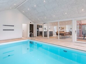 A modern indoor swimming pool with clear glass walls, a kitchen area, and a dining space. - 10 Person Holiday Home in Jerup (Jerup)