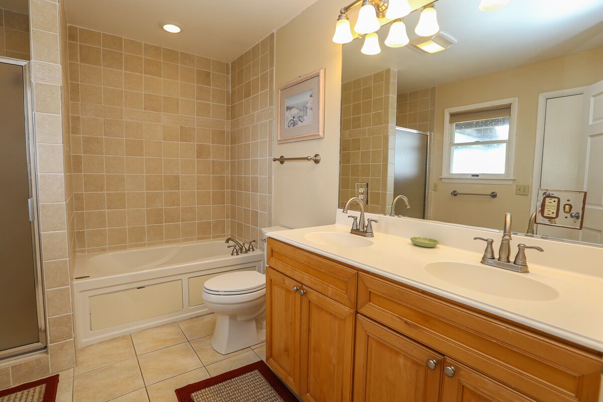Une salle de bain avec des carreaux de couleur beige, une baignoire, des toilettes, un lavabo avec des meubles en bois et un miroir.