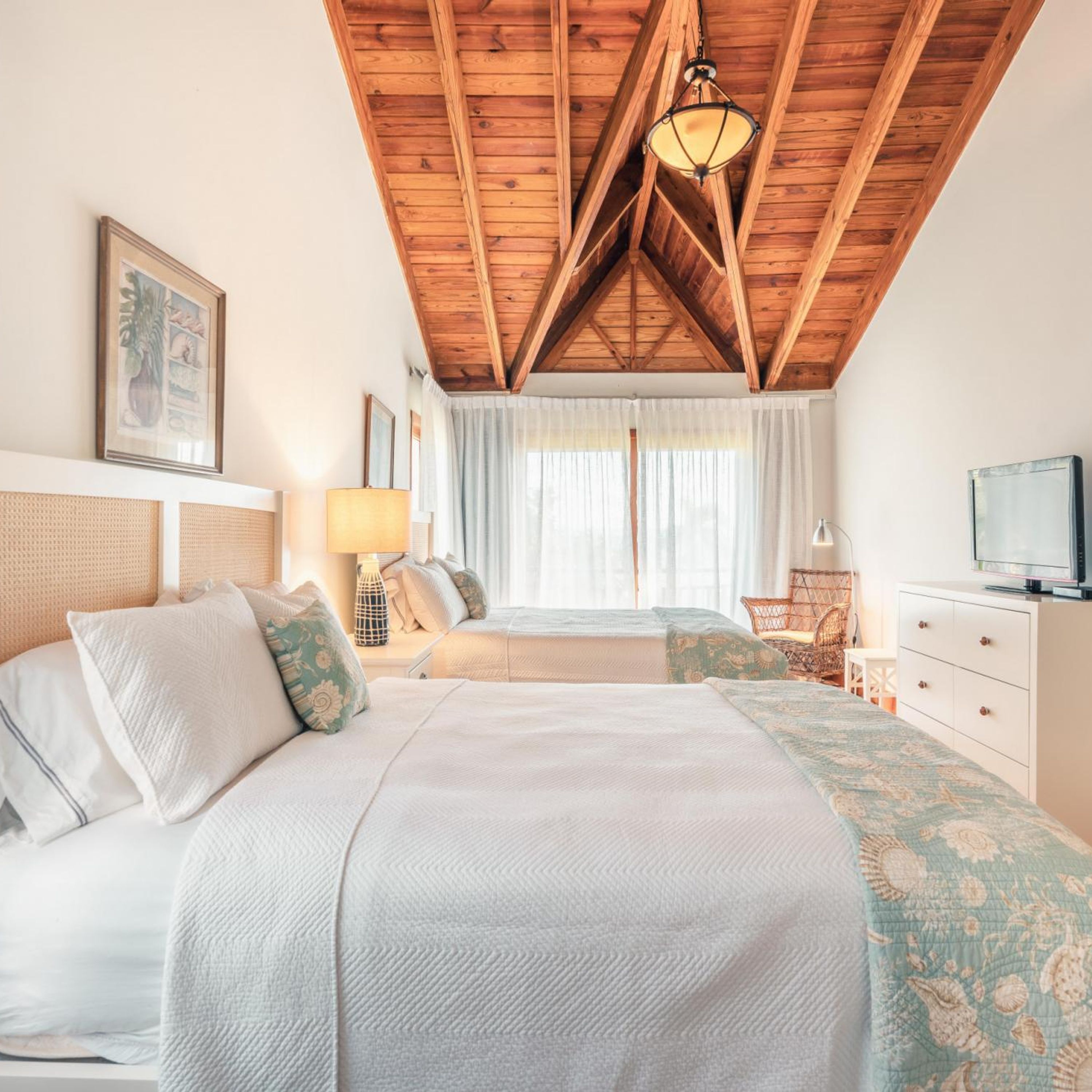 A bedroom with a wooden ceiling, a bed with white bedding, a bedside table with a lamp, a chair, and a television on a dresser.