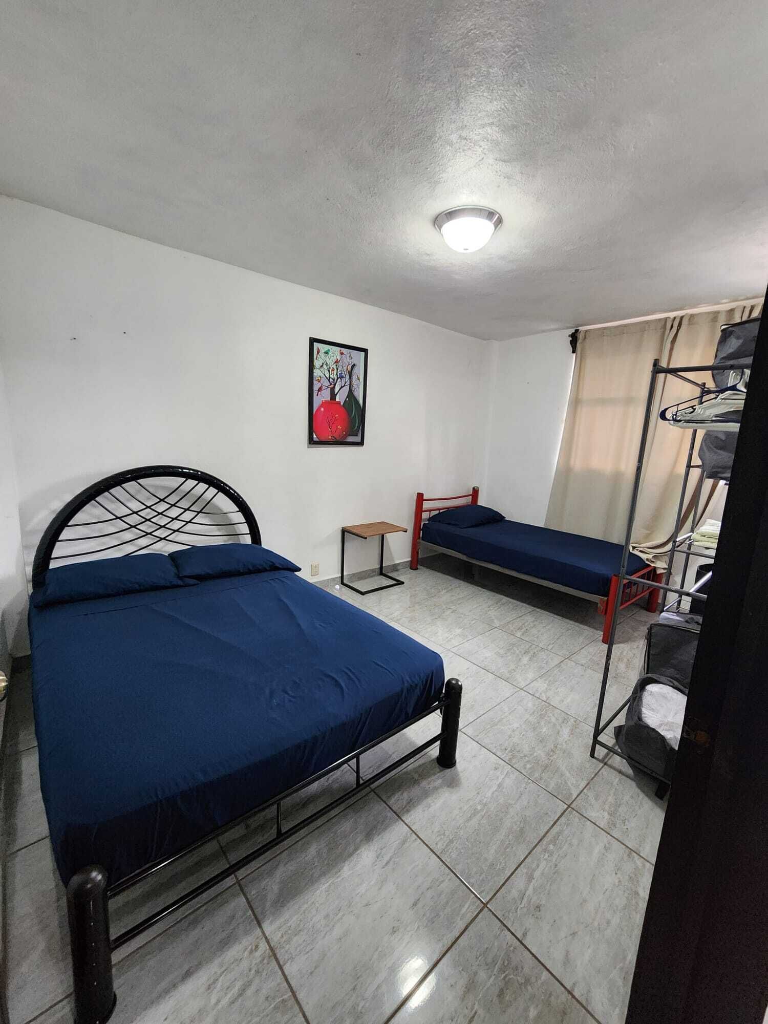 Une chambre de dortoir avec deux lits simples, une petite table, une étagère avec des vêtements pliés et un tableau accroché au mur.