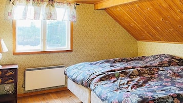 A bedroom with a wooden ceiling, a window with floral curtains, a bed with a floral bedspread, a bedside table, and a radiator.