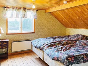 A bedroom with a wooden ceiling, a window with floral curtains, a bed with a floral bedspread, a bedside table, and a radiator. - 5 Person Holiday Home in Fargelanda (Färgelanda)
