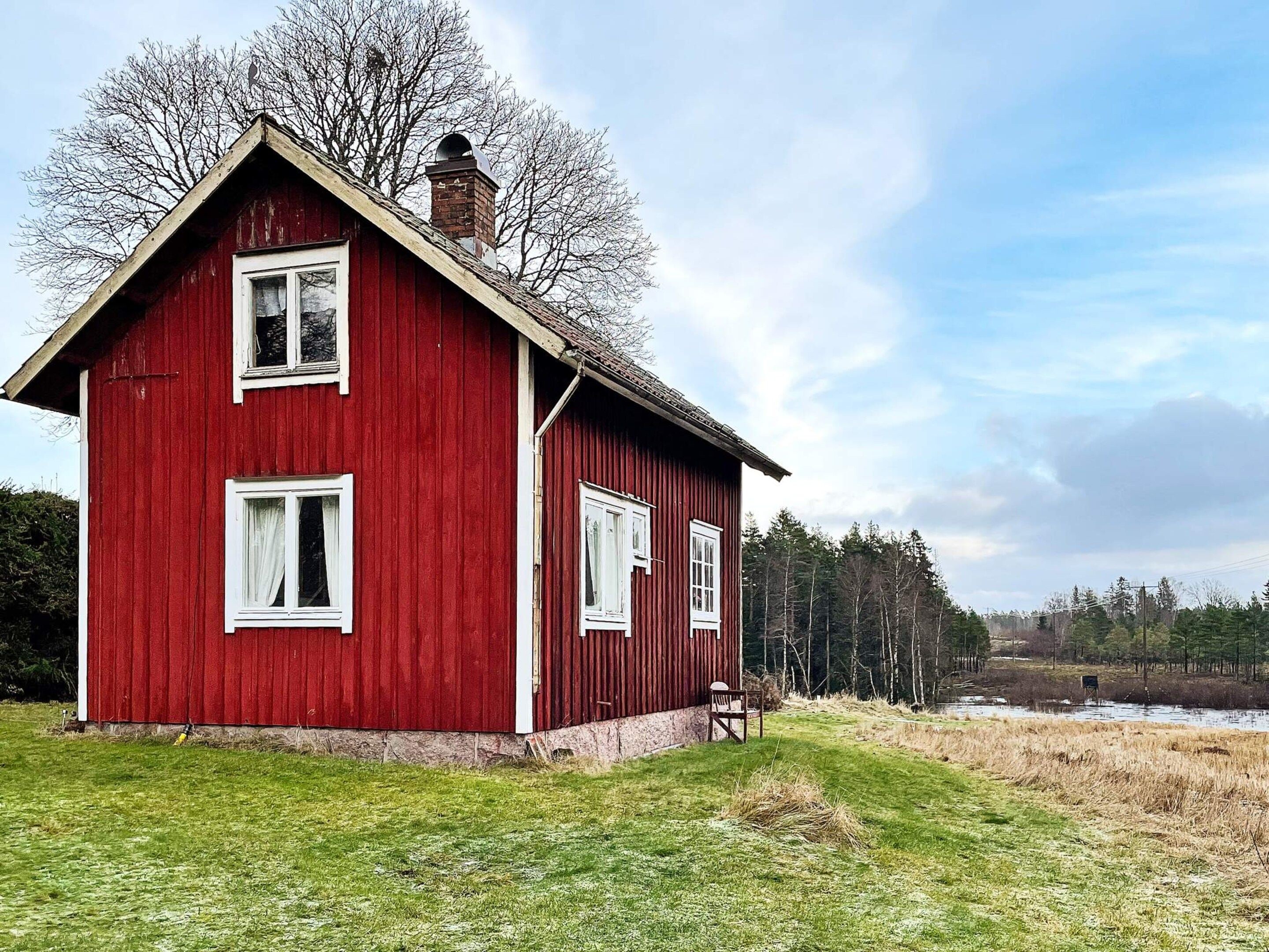 A red wooden house with two windows and a chimney, surrounded by a grassy field and trees.