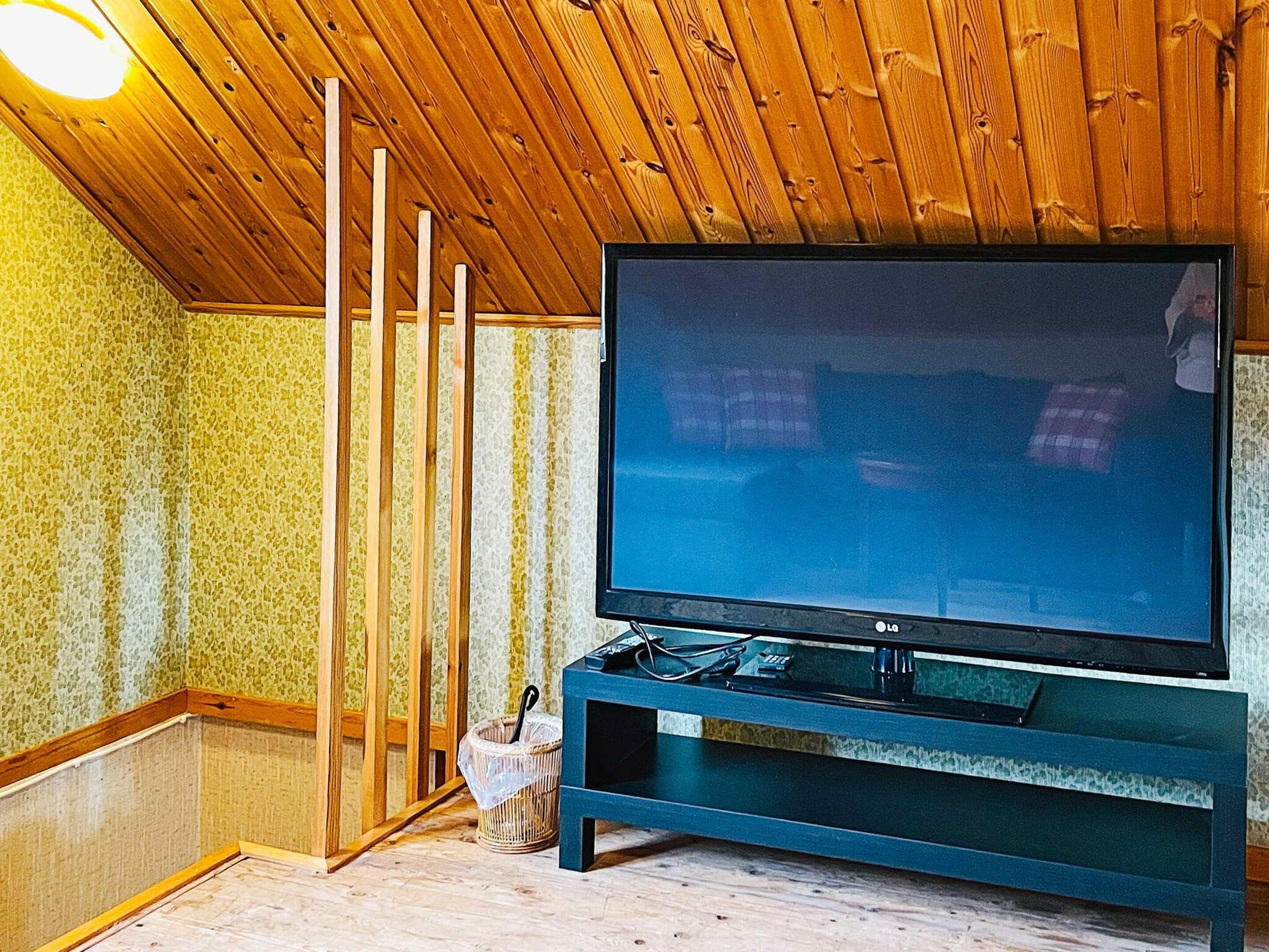 A room with a large flat-screen TV on a stand, wooden ceiling, and patterned wallpaper.