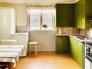 Een kleine keuken met houten kastjes, een krukje, een raam met bloemen gordijnen, en een koelkast.