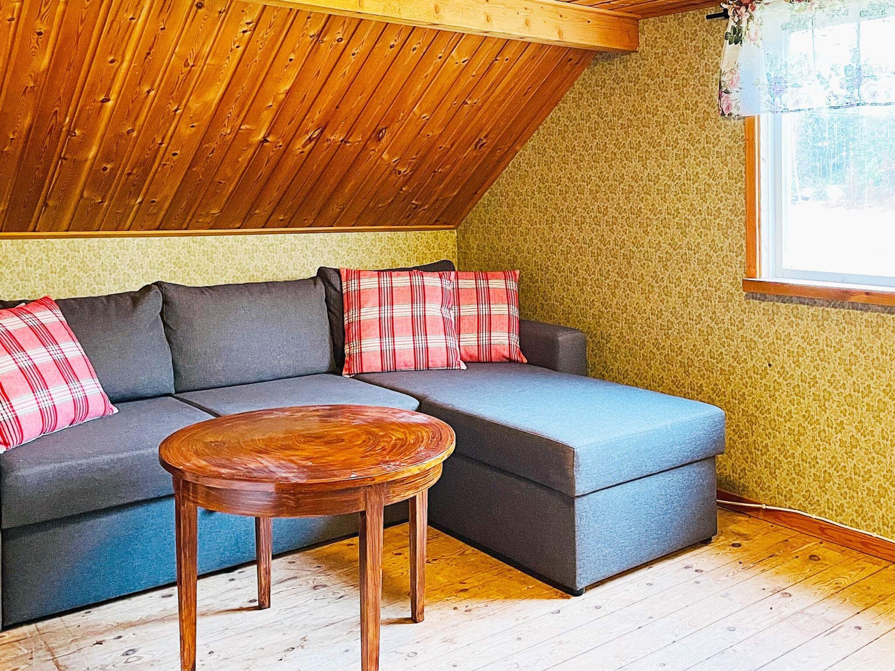 A cozy room with a sloped ceiling, a gray sectional sofa with plaid cushions, a wooden coffee table, and a window with a floral curtain.