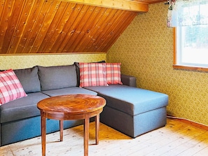 Une pièce chaleureuse avec un plafond en pente, un canapé d’angle gris agrémenté de coussins à carreaux, une table basse en bois et une fenêtre ornée d’un rideau à motifs floraux.