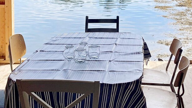 Una mesa para dos con mantel a rayas, dos sillas y una jarrón de vidrio sobre la mesa.