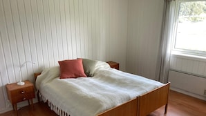 Un dormitorio con una cama de madera, dos mesitas de noche, una mesita auxiliar, una ventana con cortinas y un radiador.