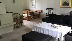Una sala de estar acogedora con un sofá negro, una mesa de madera con mantel blanco, un banco de madera, un rueca y un cuadro en la pared.