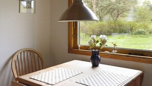 Een kamer met een tafel, een stoel, een raam met uitzicht op groen, een hanglamp en een vaas met bloemen.