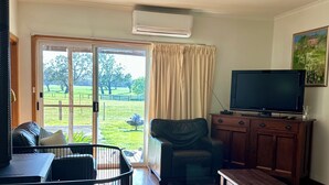 A living room with a wooden floor, a black leather sofa, a wooden coffee table, a television on a wooden cabinet, and a sliding glass door leading to a garden. - A charming cottage with picturesque rural views over surrounding rural farm land (Melville Forest)