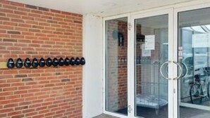 Een bakstenen muur met een rij zwarte voorwerpen, een glazen deur met een handgreep en een glimp van een fiets van binnen.