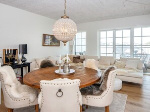 A spacious living room with a round wooden dining table, white upholstered chairs, a chandelier, and a large window providing a view of the outdoors.