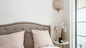 Een bed met een gestoffeerd hoofdbord, een nachtkastje met een lamp, een vaas met bloemen en een kaars.