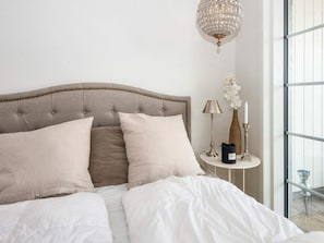 Uma cama com cabeceira estofada, uma mesa de cabeceira com abajur, um vaso com flores e uma vela.