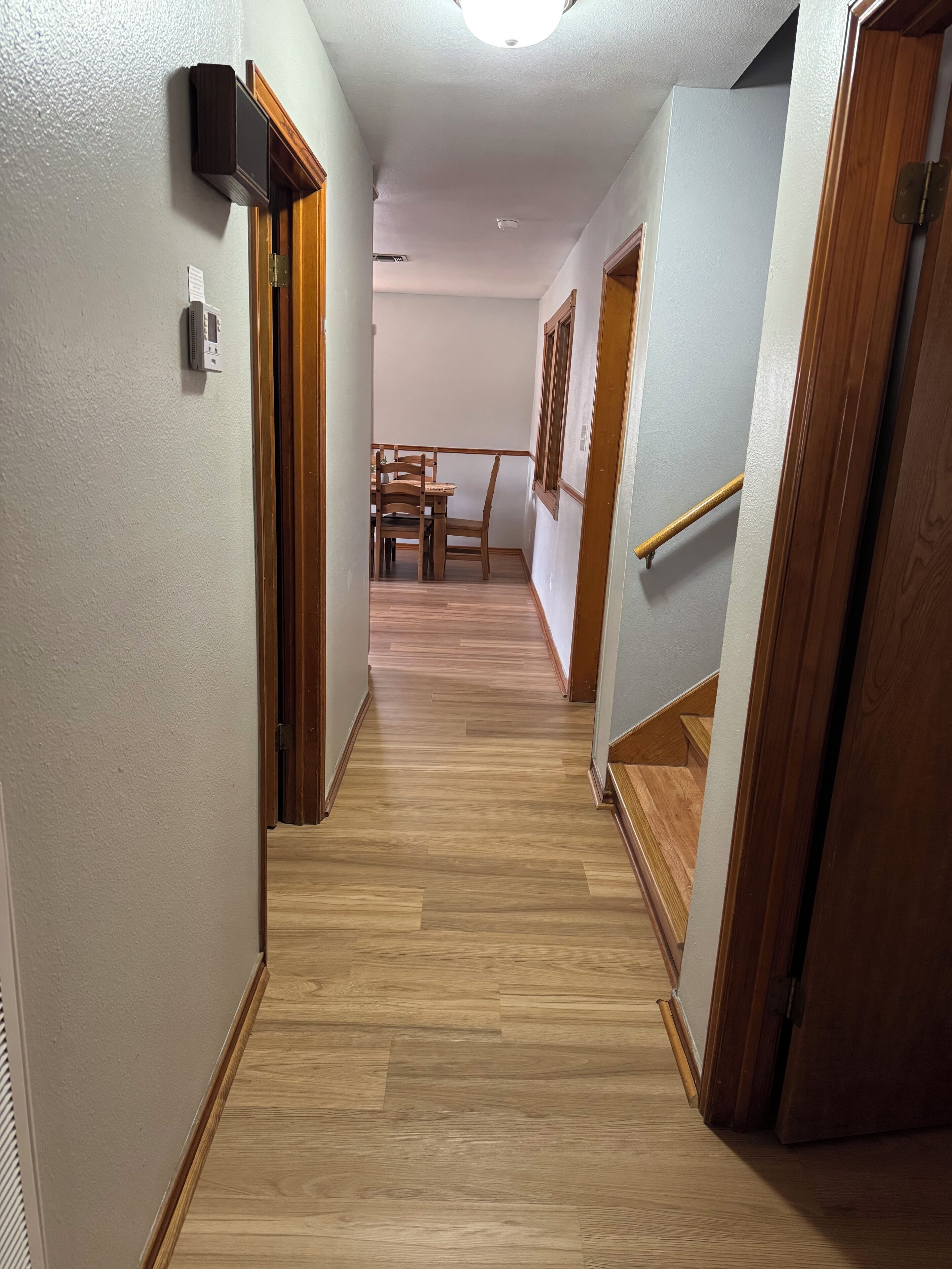 A narrow hallway with wooden flooring, a dining area with chairs and a table, and a staircase with wooden railings.