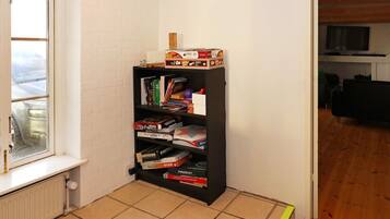 Habitación esquinera con una estantería llena de libros y juegos de mesa, un radiador y una ventana con vista a un coche estacionado.