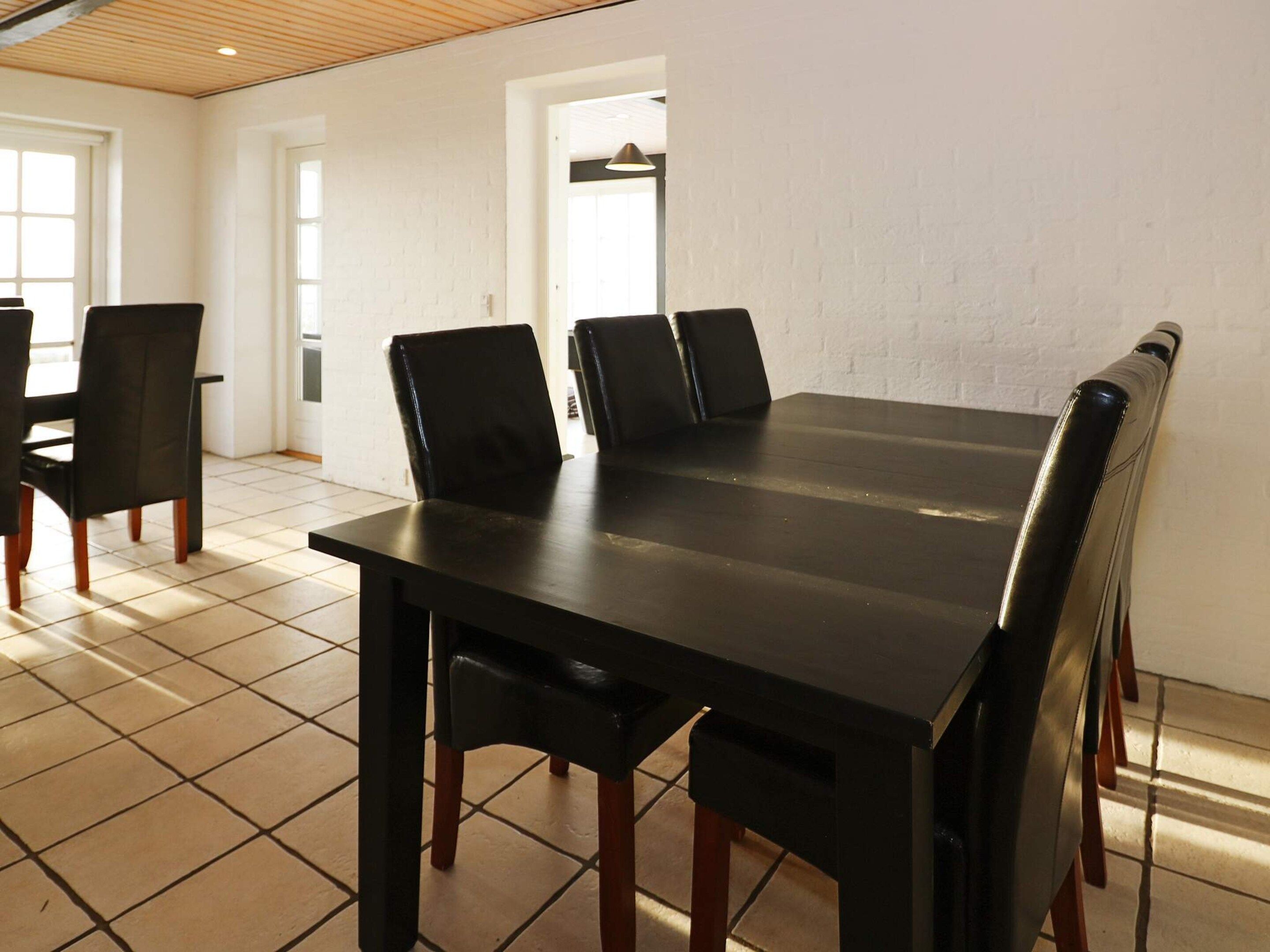 A dining area with a long table and chairs, white brick walls, and tiled flooring.