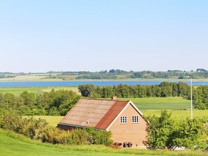 A single-story brick house with a sloped roof, surrounded by lush greenery and a body of water in the background. - 14 Person Holiday Home in Havndal (Havndal)