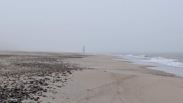 Uma praia com pedras e areia, uma pessoa em distância e um farol ao fundo.