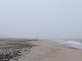 A beach with pebbles and sand, a person standing in the distance, and a lighthouse in the background.