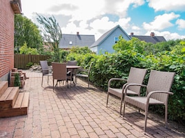 A patio with wicker furniture and a brick floor, surrounded by greenery and a wooden fence.