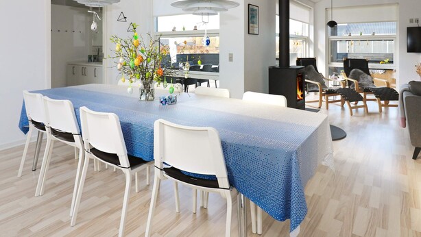 A modern dining area with a long table, white chairs, and a blue tablecloth. There is a black stove in the background, and the room has white walls and wooden flooring. - 8 Person Holiday Home in Hadsund (Hadsund)