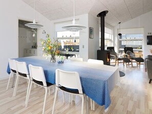 A modern dining area with a long table, white chairs, and a blue tablecloth. There is a black stove in the background, and the room has white walls and wooden flooring. - 8 Person Holiday Home in Hadsund (Hadsund)