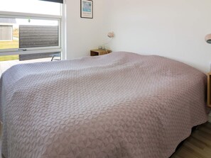 A bedroom with a large bed, a small bedside table, a wall-mounted lamp, and a window with a view of the outside.