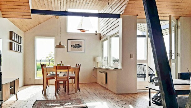 Une pièce spacieuse avec un plafond haut, des poutres en bois, un coin repas avec une table et des chaises, une fenêtre donnant sur la verdure et un grand miroir.
