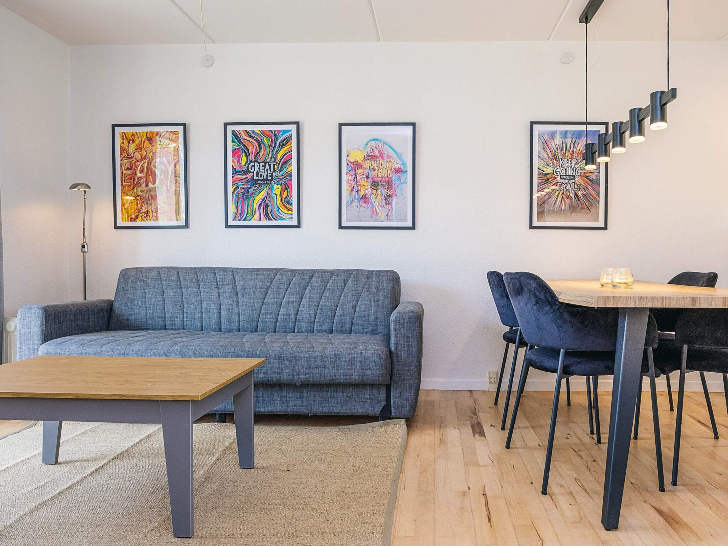 A modern living room with a grey sofa, a wooden coffee table, and a dining area with a wooden table and black chairs. There are three colorful abstract paintings on the wall.