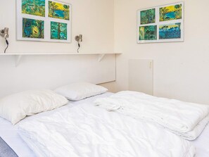 Una cama con ropa blanca, una pared blanca y un estante con fotografías enmarcadas.
