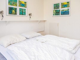 Una cama con ropa blanca, una pared blanca y un estante con fotografías enmarcadas.