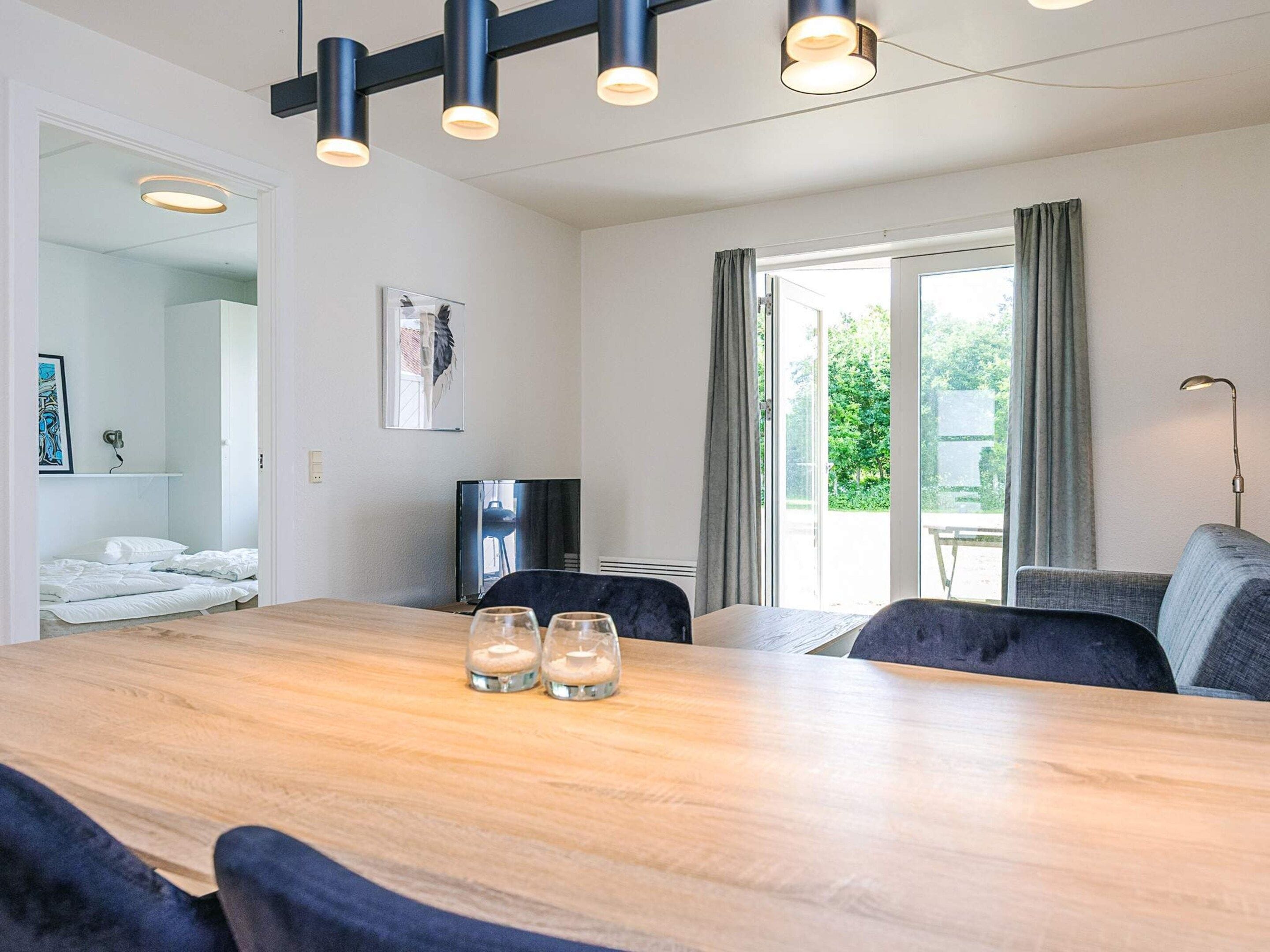 A modern room with a wooden dining table, two chairs, a sofa, a TV, and a large window with curtains.